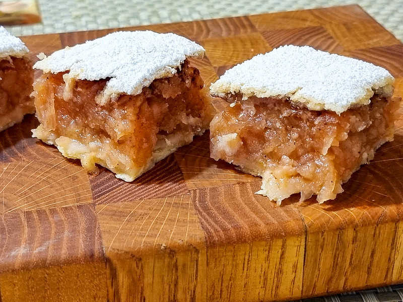 Two square slices of apple pie on a wooden chopping board. You can see their section, which is full of grated and cooked apples. On the top shortcrust there is icing sugar.