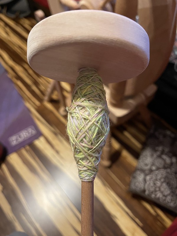 a pale wood top-whorl drop spindle with pastel pink / green / yellow singles spun, and wrapped on it in a criss-cross pattern
