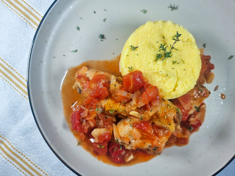 A grey plate with the chicken stew on it near a round mamaliga. The stew is red from the tomatoes.