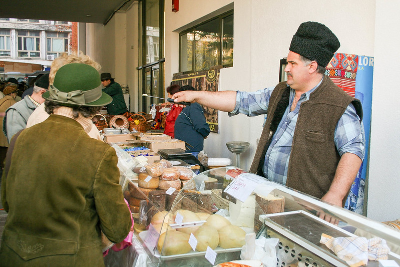 A man in the market offering a potential customer a small piece of cheese, to try it on.