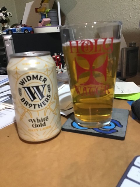 Widmer's White Gold hazy lager can, next to a glass of same on a messy desk