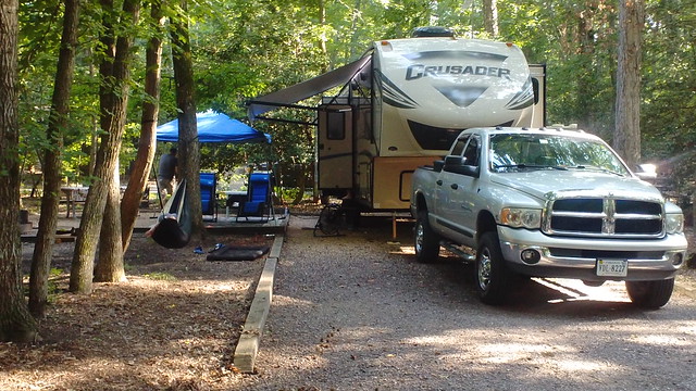 truck and RV set up at campsite with a blue pop up tent next to it, with blue recliner chairs and a person laying in a hammock strung between two trees among many trees surrounding the site