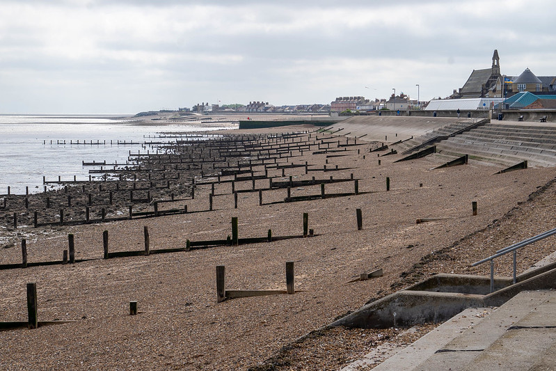 A pebbles beach divided by many wooden poles at equal distances from each other. The water is on the left, and the tide is low.