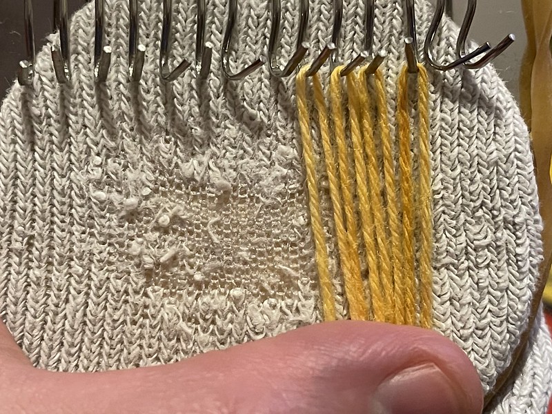 metal hooks of a darning loom on a white commercial sock with yellow sock-weight yarn being warped to start creating a woven darn repair