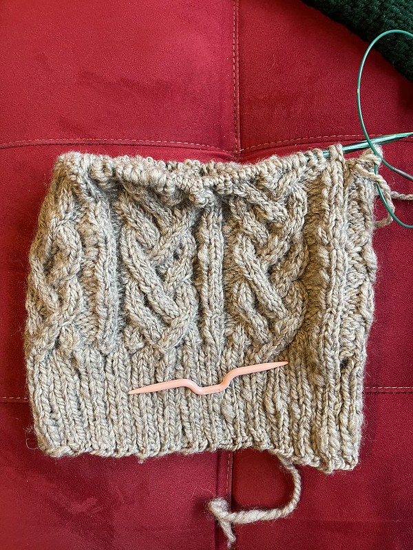 several inches of an in-progress cabled hat on green wooden circular knitting needles with a pink cable needle laying on top; the yarn is a sandy grey wool hand spun yarn that varies in weight from about sock weight to a heavy worsted weight; in spite of the variable weight of the yarn, the cables pop in a visually noticeable way