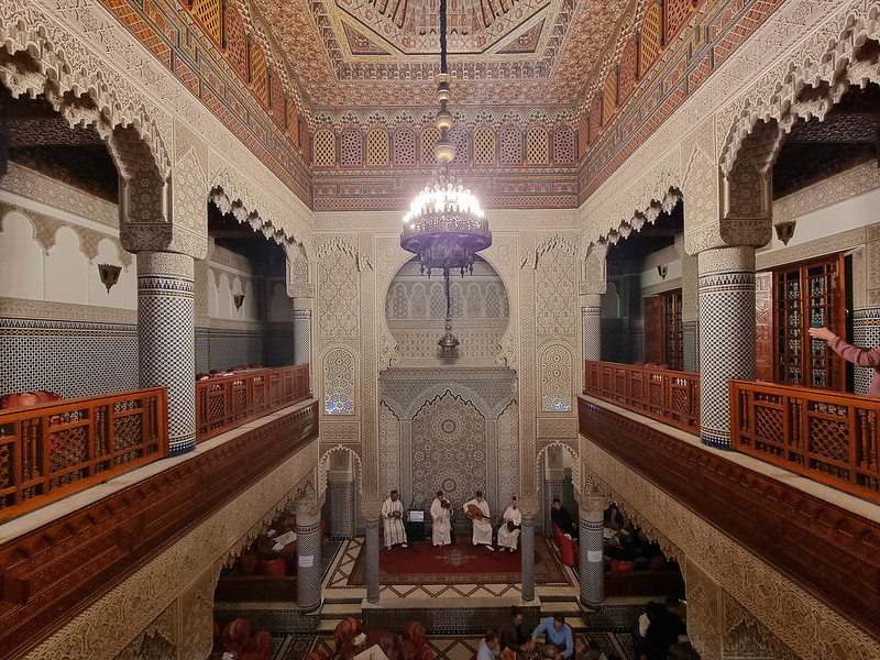 The interior of a Moroccan Riad, with a covered interior courtyard which is now a restaurant. The walls are covered in mosaics and there is a large chandelier hanging from the ceiling. At the bottom, right in front, there are four men wearing white costumes, playing traditional Moroccan instruments.