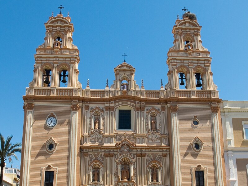 The facade of the Cathedral in Huelva. It has two large bell towers on each side, and a smaller one in the middle. There are different statues on the facade, in the middle part