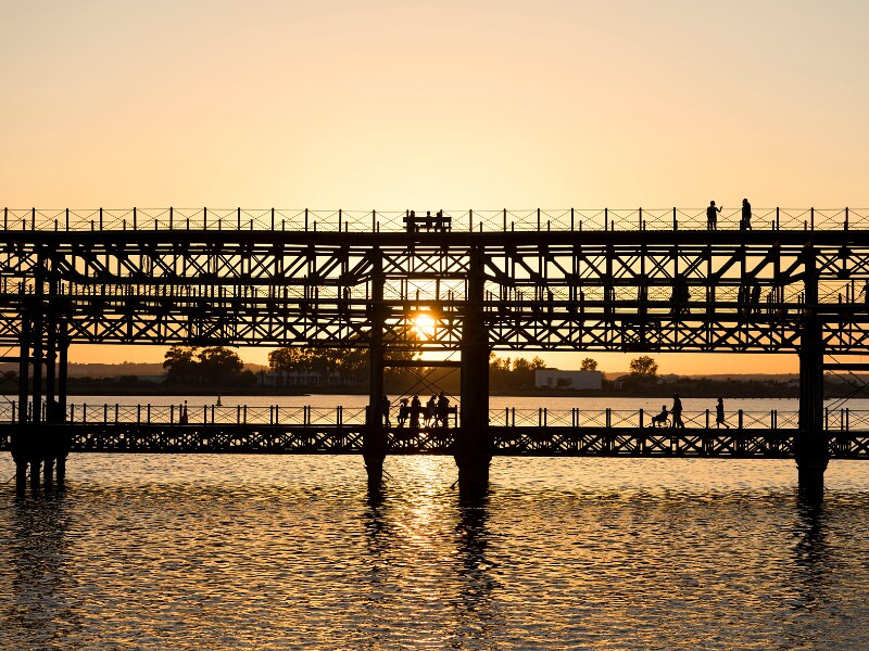An iron pier on two levels, with people walking on both, at sunset. The ski is orange and the sun can be seen through the iron work of the pier.