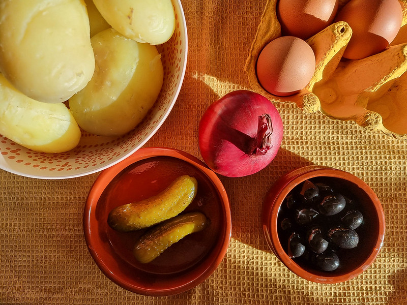 A white bowl with pink polka dots on the left hand side of the photo, holding boiled, peel potatoes in it. On the right, there is a carton with three eggs in it. Below, there is a ceramic terracotta ramekin with two pickles in it, another one filled with olives, and a red onion between them. They are sitting on a yellow tea towel.
