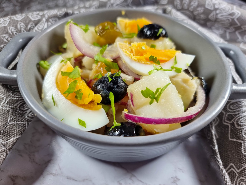 A close-up of the potato salad, inside a grey ramekin with two handles. You can see the potatoes, slices of boiled eggs, black olives cut in half, and slices of red onions.