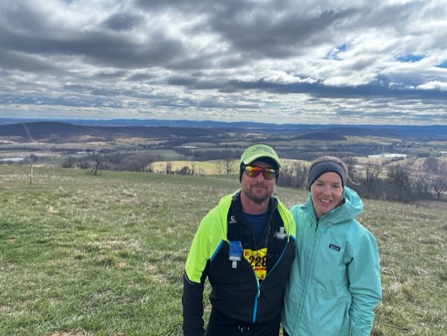 smiling man and woman in cold-weather athletic attire standing before a scenic vista of rolling hills.