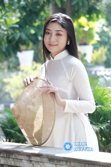 A charming, beautiful girl with silky black hair is posing gracefully in an Ao Dai, the white traditional Vietnamese dress, and an Asian conical hat in the temple courtyard.