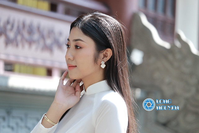A charming, beautiful girl with silky black hair is posing gracefully in an Ao Dai, the white traditional Vietnamese dress in the temple courtyard.