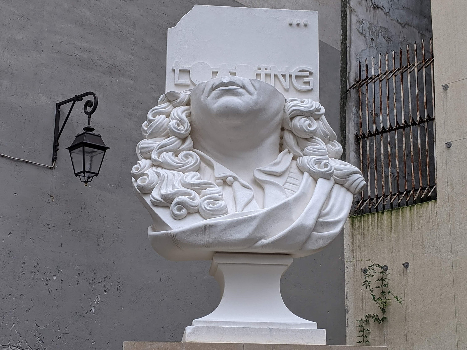 A classical white bust stands on a pedestal in front of a wall (in Paris). The top portion of the bust is replaced by the top of the document ison from a computer, with the word "LOADING" written on it