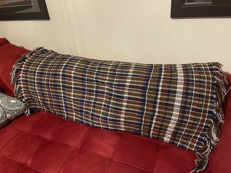 A full length of fabric (a bit less than 4 feet) draped over the back of a red couch. The plaid patterning is irregular, with most of the white bands narrower than the red, olive, and blue stripes, but for one thicker white stripe towards the right. Both ends of the fabric have fringe 