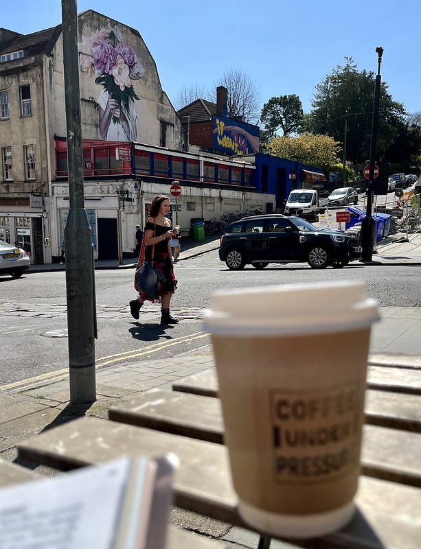 A photo from the point of view of sitting at a table outside a coffee shop, looking across Park Street in Bristol, on a sunny day. An out of focus disposable coffee cup and a magazine are in the foreground. A young woman wearing big lace-up boots, a long skirt and an off-the-shoulder top is walking past. A mural high up on the building opposite depicts someone holding a large bunch of purple and orange flowers in front of their face.