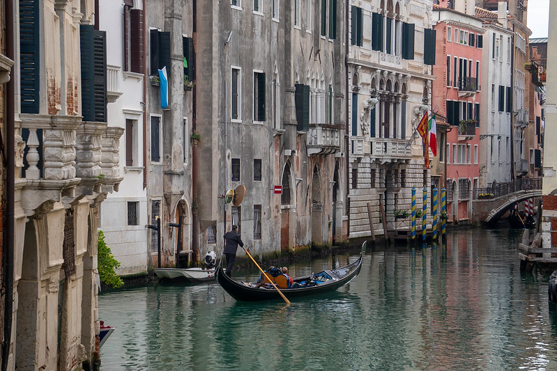 A lone gondolier rowing his two clients on an empty canal. The houses alongside the canal are all different, some very simple, some with decorations and bright facades.