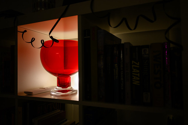 Crimson glass goblet illuminated inside a backlit bookcase cube, surrounded by books and decorative curling wires overhead.