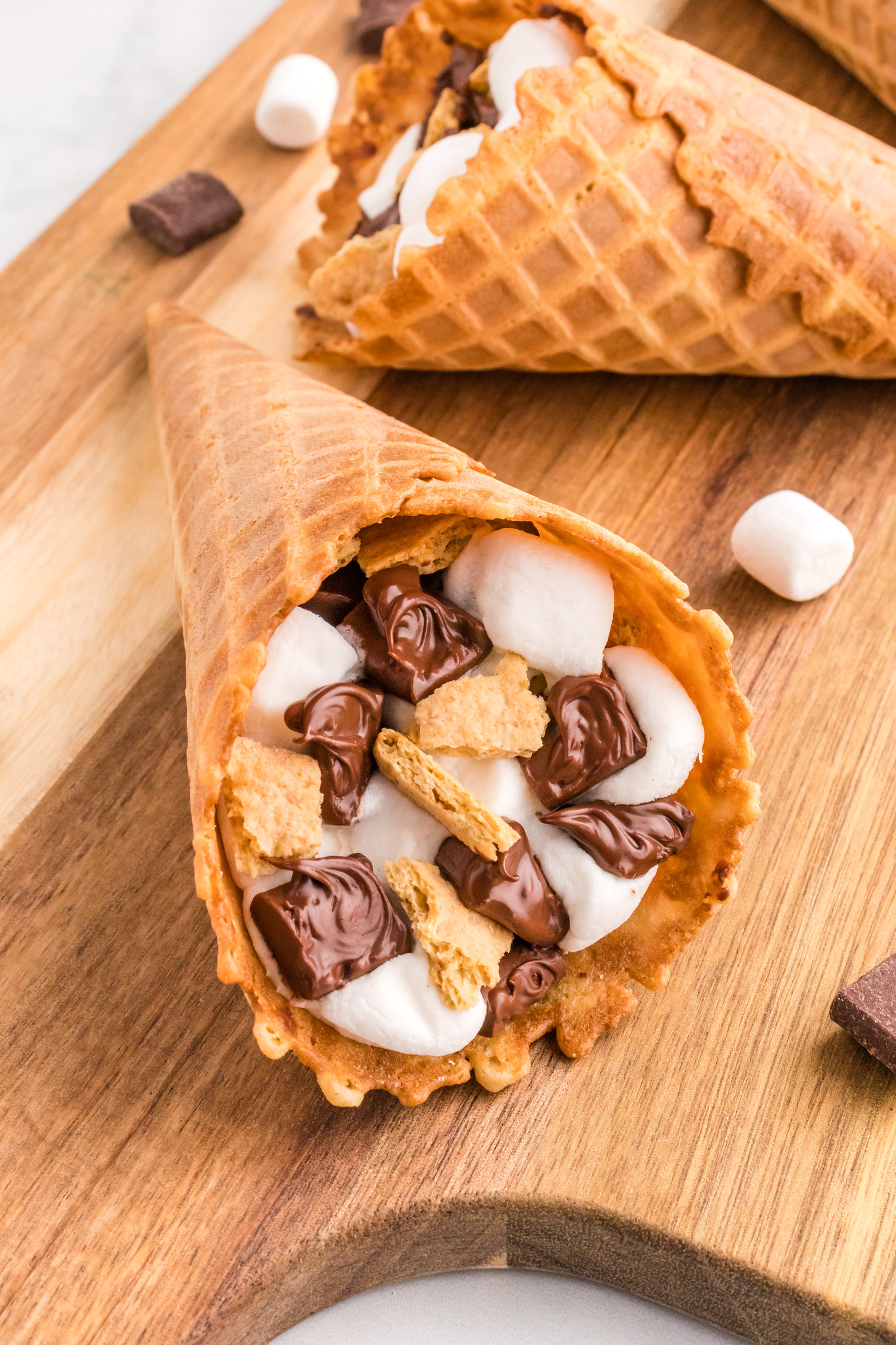 S'mores Campfire Cone filled with marshmallows, chocolate, and graham cracker pieces. On a wooden serving tray with marshmallows and chocolate scattered around. 