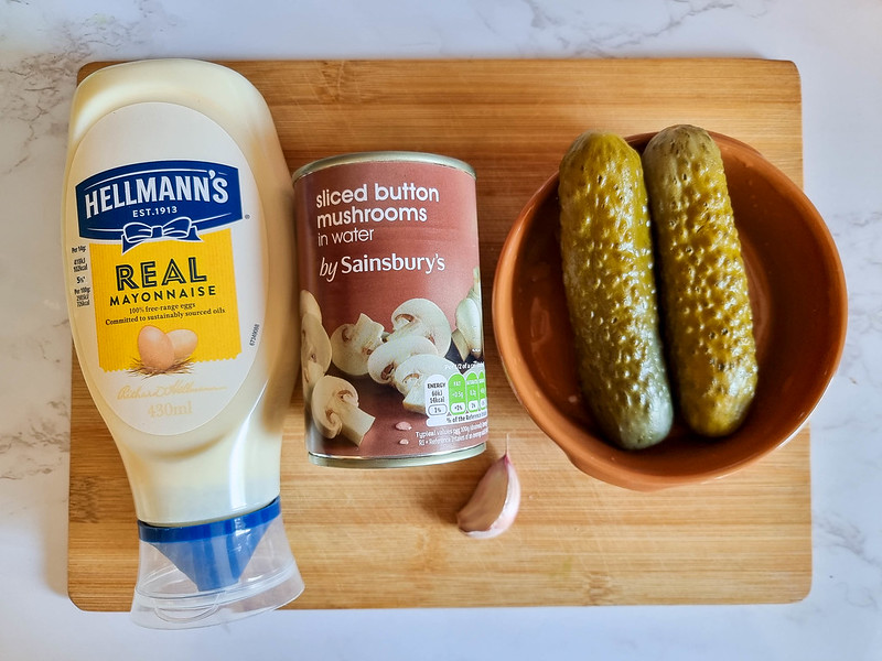 A tub of Hellmann's mayo, a can of sliced mushrooms, a garlic clove and a ramekin with two pickles, all on a wooden chopping board.