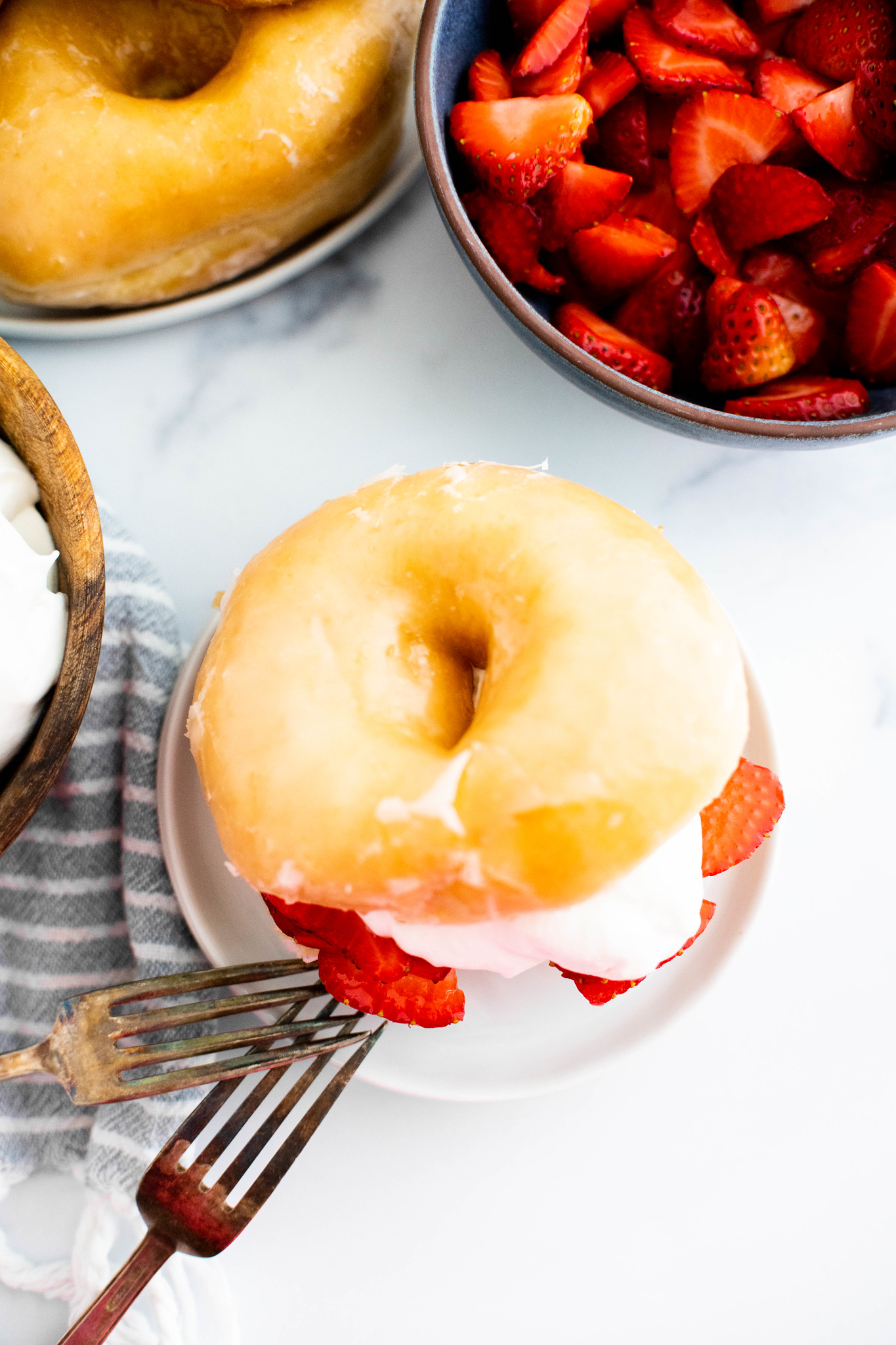 Glazed donut sliced in half filled with macerated strawberries and homemade whipped cream. On a white round plate with a bowl of strawberries in the background and a tower of glazed donuts.