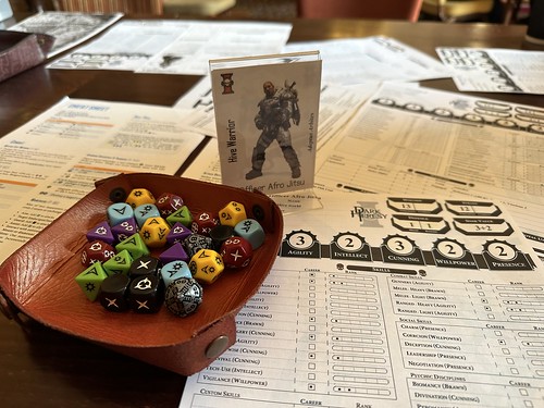 A game of WH40K hacked to Genesys at North Star 2023. The photo shows a character sheet, a dice tray with multi-coloured Genesys dice and a character standee entitled 'Hive Warrior'.