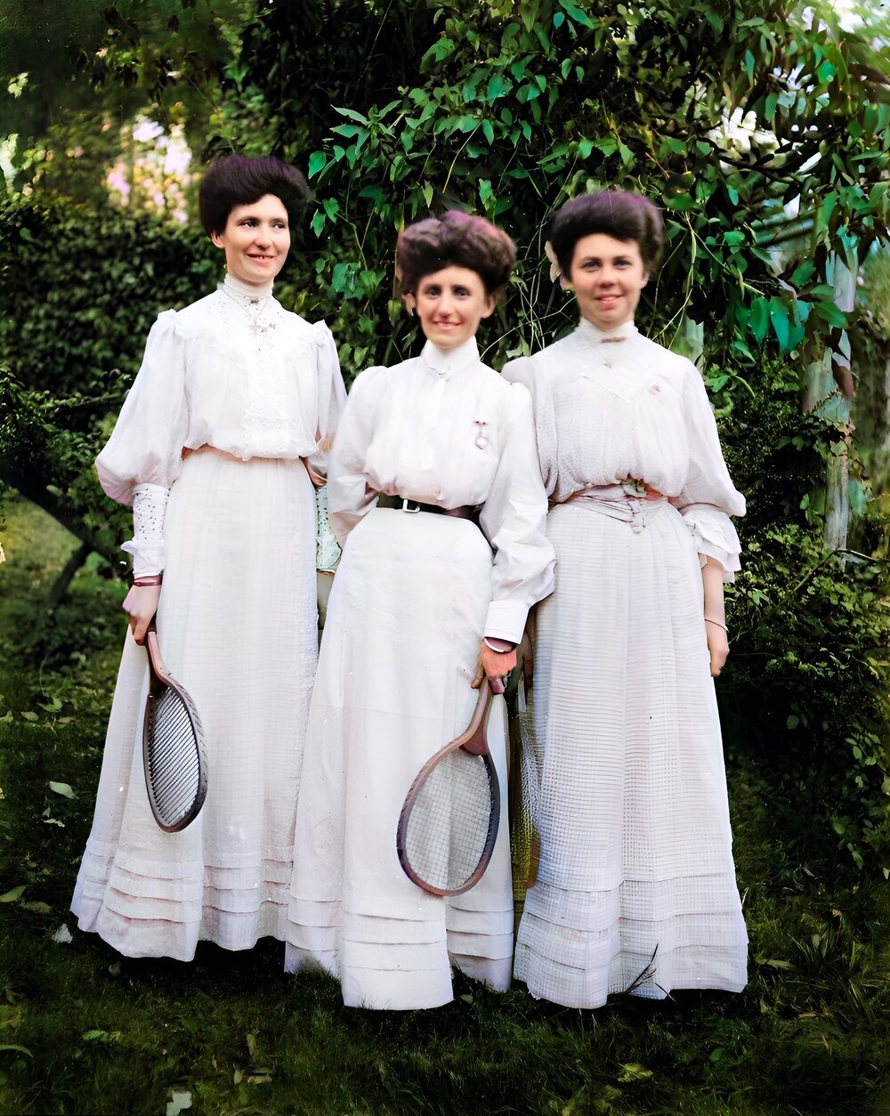 Three young women in light dresses holding tennis racquets, 1900