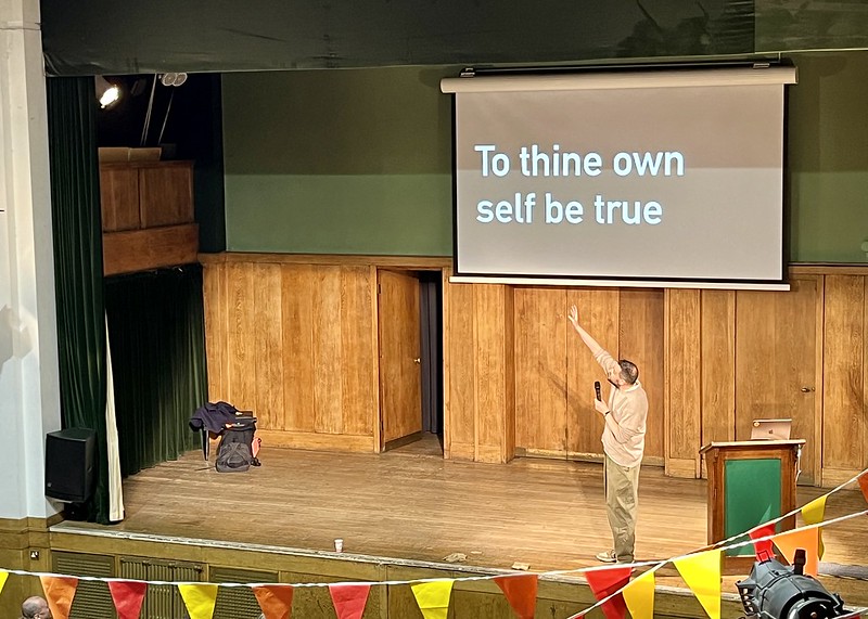 A photo of a wooden stage with Russell gesturing at a big screen on which text reads 'To thine own self be true'. In the foreground is some red and yellow bunting.