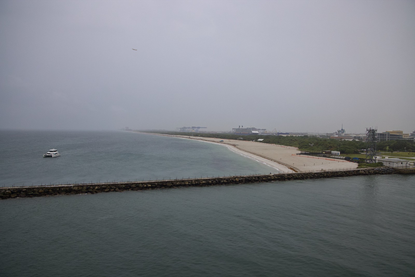 Our ship leaves port, passing the breakwater and beach in the rain as an airliner climbs out from the airport