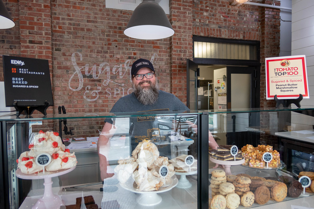 Jeff Nachtigall stands behind a glass case full of baked goods with a brick behind him emblazoned with the Sugared and Spiced logo