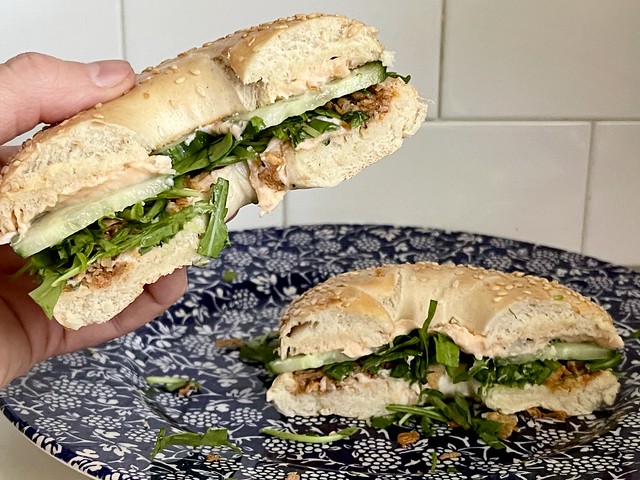 Savoury and sweet smoked salmon spread in a sesame bagel sandwich with rocket/arugula, cucumber, fried onions, and love. It is sliced in half with one half on a navy and white plate and the other half held in the foreground ready to be enjoyed. White subway tile is in the background.