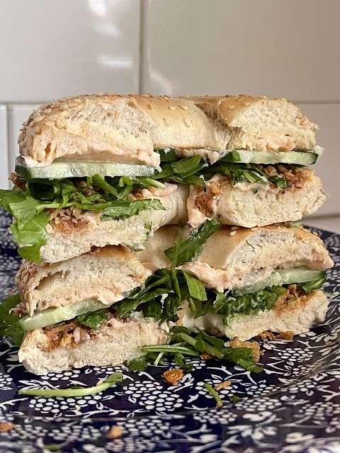 A sesame bagel with smoked salmon spread, sliced cucumber, rocket/arugula, fried onions, and love on a navy and white plate with white subway tile in the background.