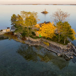 Majestic Yellow Tree and Two Humble Islands This aerial photograph captures the serene beauty of two humble islands in the heart of the 1000 Islands region, a popular destination for adventure travel in Ontario, Canada. The foreground island, shaped like a triangle, is adorned with a mix of green foliage and one strikingly majestic yellow tree, a beacon of autumn colors amongst its verdant neighbors. The island also features a quaint cottage, providing a glimpse of the simple, waterfront living that epitomizes cottage life in this area.

A calm river embraces the islands, its tranquil waters reflecting the vibrant scenery above. A small dock extends from the foreground island, inviting watercraft travelers to explore the natural splendor that surrounds them. The background island, even smaller in size, adds depth to the composition and underscores the sheer number of islands that make up this breathtaking Canadian landscape.

The photograph, skillfully taken by Duncan Rawlinson, reveals the allure of the 1000 Islands and the enchanting outdoor living it offers. The image captures the essence of the region, from its rocky shoreline to the colorful trees that mark the arrival of fall. The golden hour light further enhances the charm of the scene, casting a warm glow on the islands and their surroundings.

In this harmonious blend of nature and human habitation, one can easily imagine the quiet life on the river – a life filled with the simple joys of outdoor adventure, coastal living, and an intimate connection to the ever-changing beauty of nature. 

&lt;a href=&quot;https://Duncan.co/majestic-yellow-tree-and-two-humble-islands&quot; rel=&quot;noreferrer nofollow&quot;&gt;Duncan.co/majestic-yellow-tree-and-two-humble-islands&lt;/a&gt;