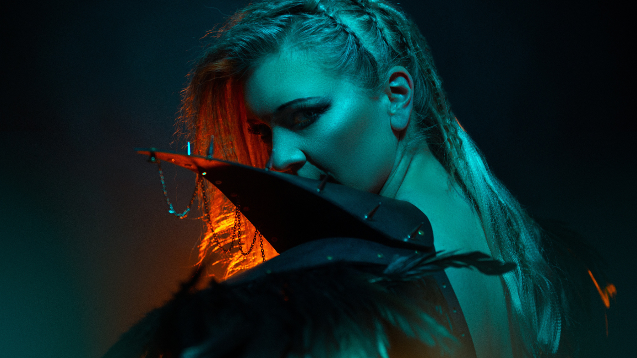 Jessi is facing the side and looking towards the camera. She has dark eyeshadow and a leather jacket. The lighting is dark, with blue and orange tones.
