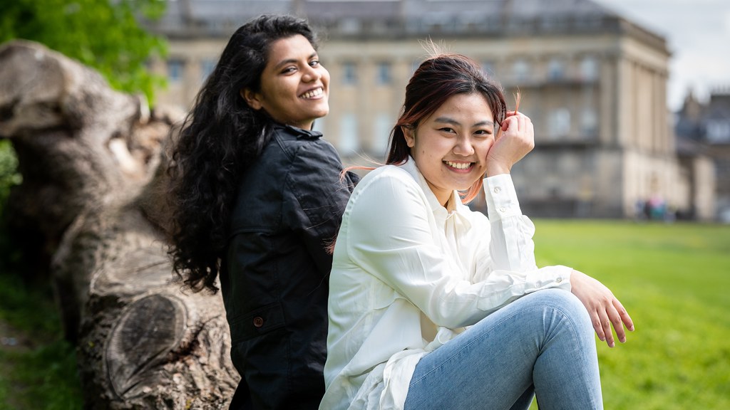Two female students sit in a park and smile towards the viewer.