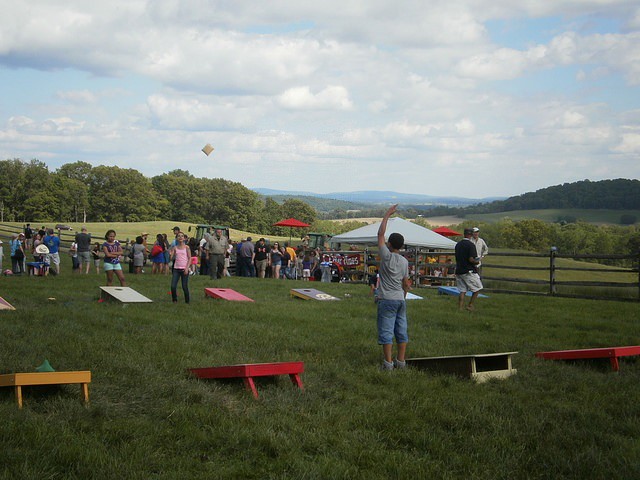 A large gathering of people on the grounds of a farm, playing corn hole, with event tents and tractors in the background.
