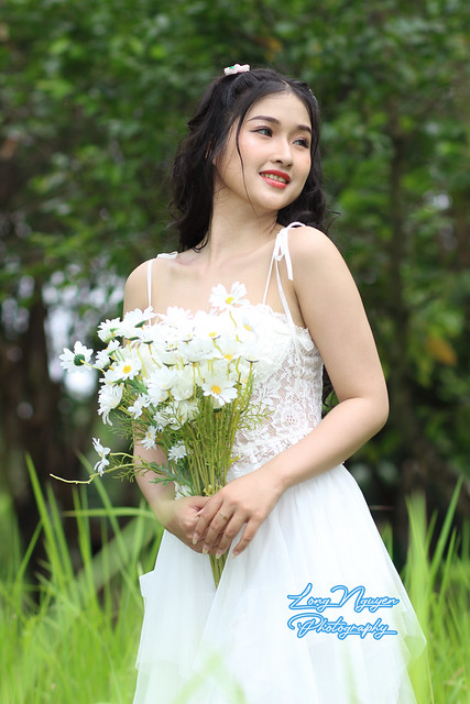 A beautiful girl with curly black hair adorned with flowers, radiating in a pure white dress, captivates with white daisies in hand, a serene moment in the heart of spring.