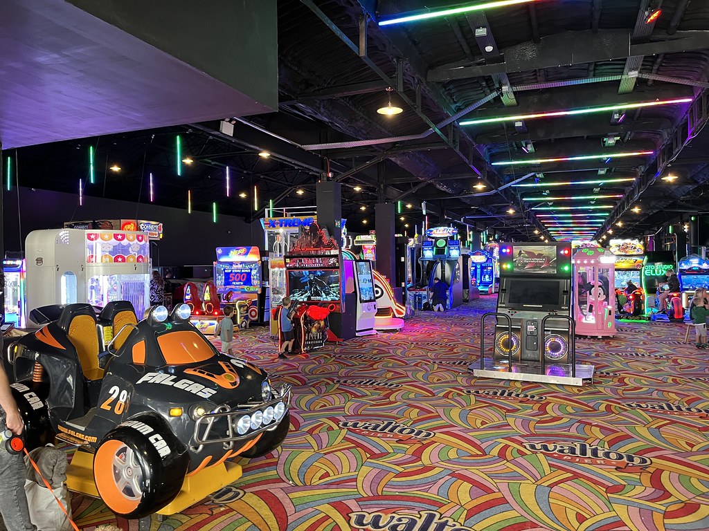 A photo of a long space stretching into the distance, with a black ceiling lit by neon tubes. The ground is covered in a swirling multi-coloured carpet. A large ride-on car amusement is in the foreground and many other amuseument machines fill the rest of the space, all lit up in different ways.