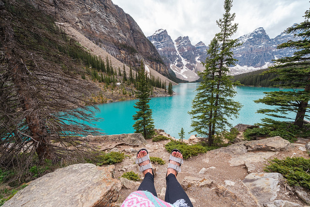 Moraine Lake in Banff National Park on an overcast day, with a woman sitting on the rocks, only feet in sandals showing