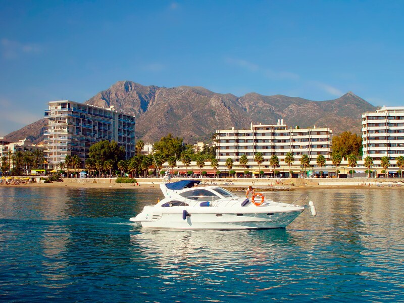 A white speed boat sailing along the coast of Marbella. The shore is very close. In the background you can see hotels, and a mountain.