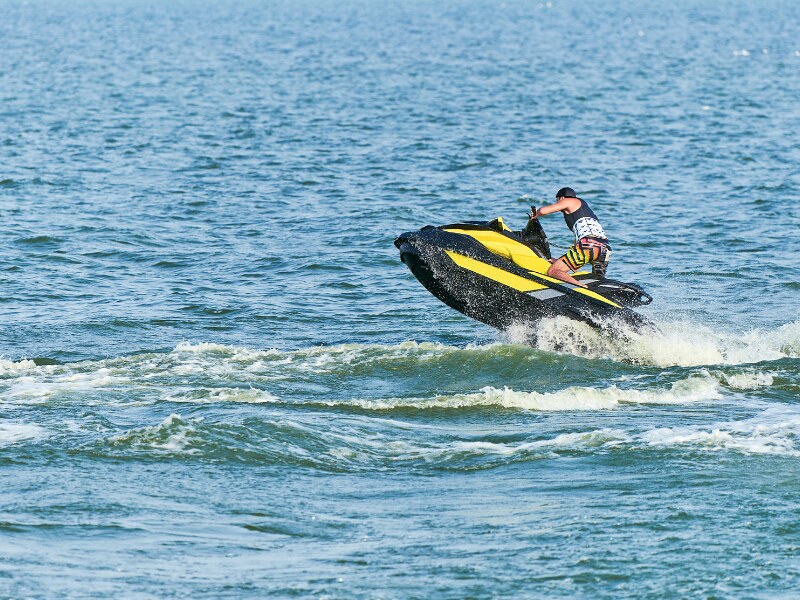 A man wearing yellow an orange shorts and a black life vest, riding a black and yellow jet ski over the water.