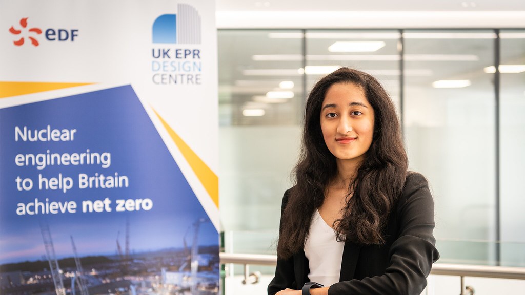 female student faces camera, smiling and standing next to an EDF banner that reads 'Nuclear engineering to help Britain achieve net zero'.