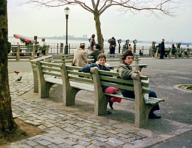 My sister and I sit on one of the benches in Battery Park in Lower Manhattan. Ellis Island can be seen in the distance as well as New York Harbor. March 1973