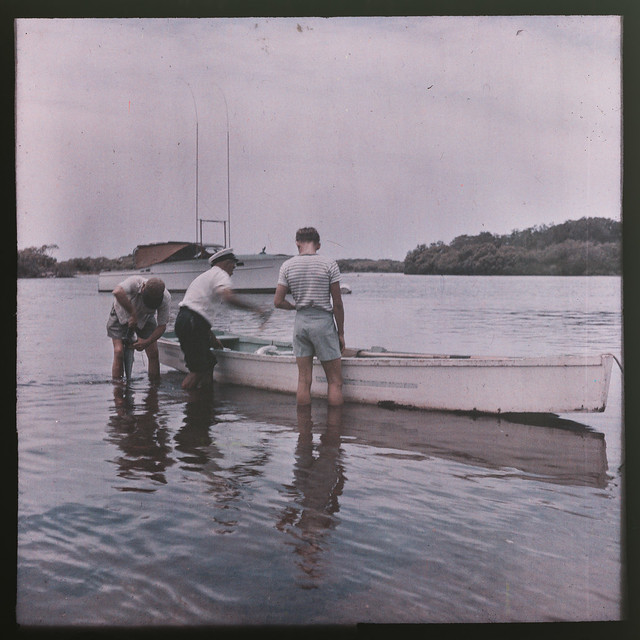 Three men standing beside a small wooden boat in shallow water, South East Queensland ca 1945