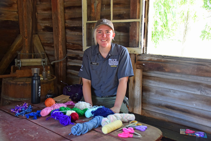 A smiling woman in AmeriCorps uniform, standing inside a rustic wood-paneled room, next to an open window and behind a table with vibrantly colored skeins of yarn on it.