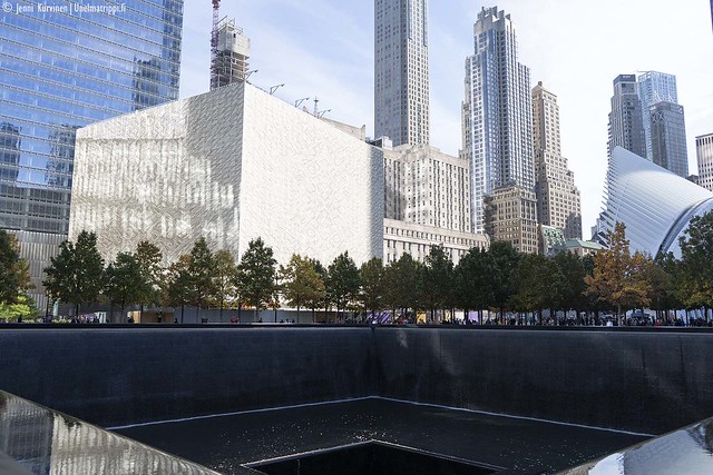 9/11 Memorialin mustaa vesiallasta, taustalla lehtipuita ja pilvenpiirtäjiä