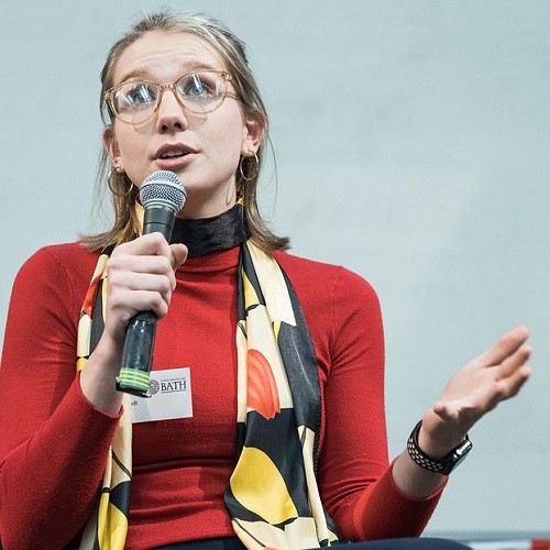 Female student wearing red jumper, black and yellow scarf speaking into a microphone to an audience behind the camera