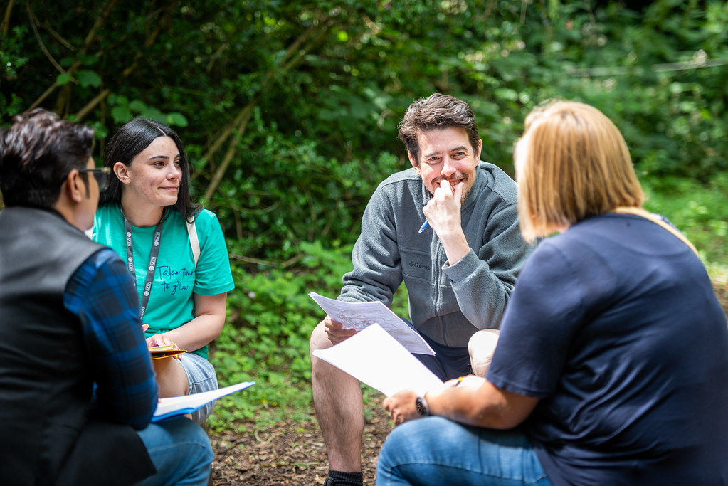 A group of people talking while sat in a sunny, wooded area.
