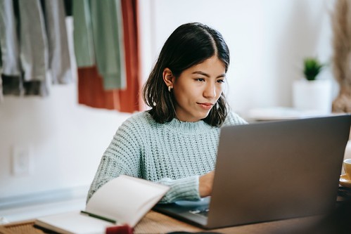 A young woman with short brown hair looks at her laptop screen. She sits at a desk wearing a light blue sweater. Next to her is an open notebook and a pen - FAFSA Help by State