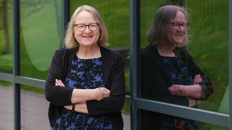 Picture of professional smart lady with glasses, standing against glass wall looking straight at the camera and smiling, you can see her reflection